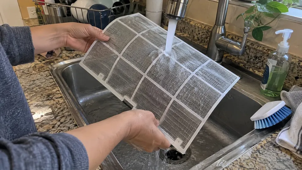 Hands rinsing a mesh mini split filter under running water in a kitchen sink with a soft brush nearby