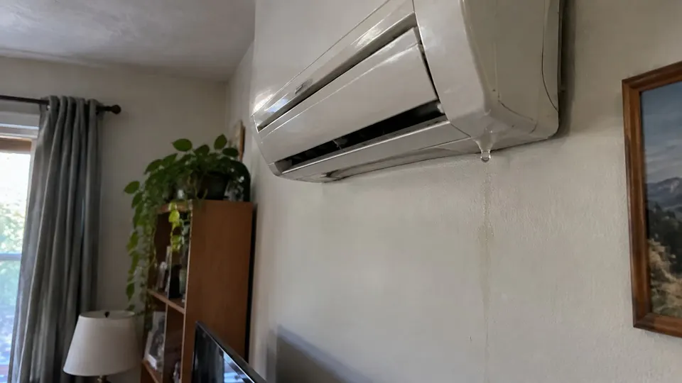 Close-up of a wall mounted mini split indoor unit with a water drop forming at its bottom edge and a faint water streak on the wall below